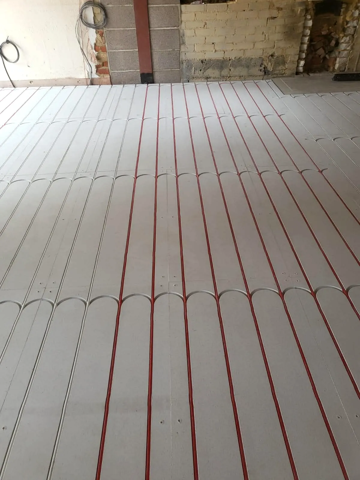 Underfloor heating manifold mounted on exposed brick wall with red pipe feeds routed across floor panels